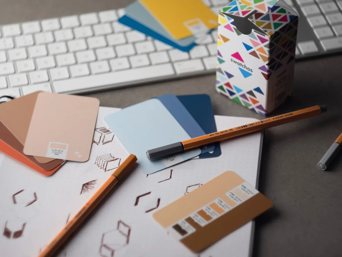 Overhead view of a designer's desk with color swatch cards, sketched icon concepts, and pencils beside a keyboard, representing GiveTurn's graphic design service